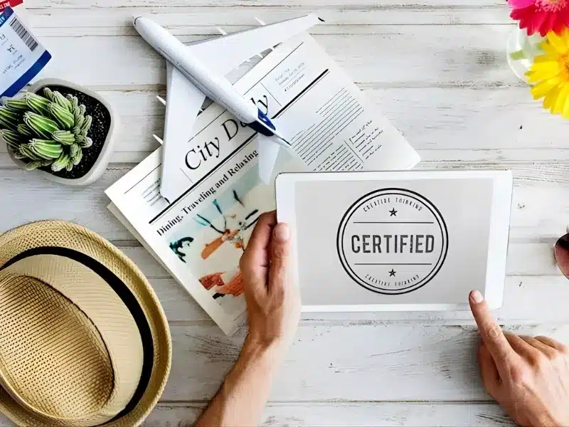 Hands holding a tablet with a certified stamp icon on a desk with travel items